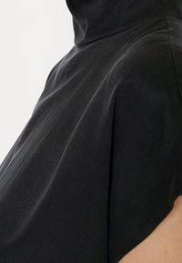 Vêtement noir ample avec un col haut et un tissu drapé. La texture semble douce, avec un design minimaliste et sans motifs visibles.