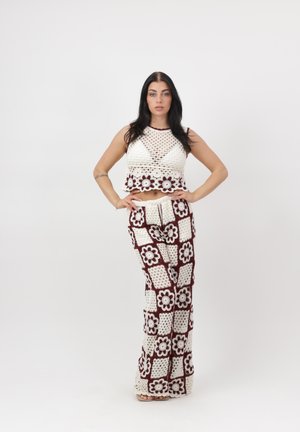 Frau steht mit Händen in der Hüfte, trägt ein weißes und burgunderfarbenes Häkeloberteil und passende weit geschnittene Hosen mit Blumenmustern.