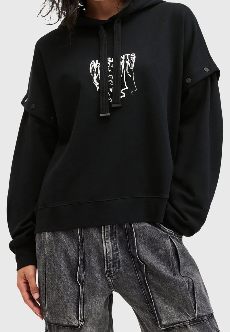 Sweat à capuche noir avec visage abstrait blanc et impression de texte, détails de boutons sur les manches, porté avec un pantalon en denim gris ample avec coutures visibles.