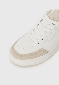 Weißer Sneaker mit einer strukturierten, cremigen Wildleder-Zehenkappe, durchbrochenem Design und auffälligen weißen Schnürsenkeln. Verfügt über ein minimalistisches Design und eine Gummisohle.