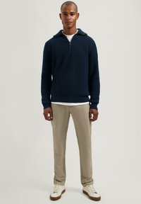 Marineblauwe geribbelde gebreide pullover met een halve ritssluiting, gecombineerd met lichtbeige broeken en witte sneakers. Zachte textuur en een strakke pasvorm.