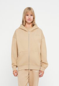 Žena v béžovej mikine na zips so sťahovacou kapucňou a zodpovedajúcich nohaviciach, stojí pred jednoduchým bielym pozadím.