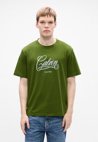 Young man wearing an olive green Calvin Klein T-shirt with white logo text, paired with blue jeans, standing against a plain white background.