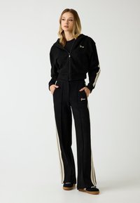 Felpa nera con zip realizzata in tessuto morbido, con dettaglio del logo. I pantaloni neri coordinati presentano righe bianche verticali e tasche anteriori.