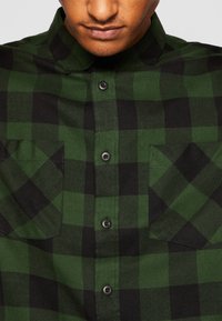 Camicia a quadri verde e nera in flanella, realizzata con un tessuto dalla texture morbida; presenta un colletto, bottoni e due tasche sul petto.