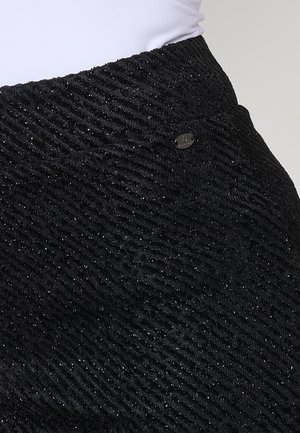 Jupe noire texturée avec un motif ondulé, des accents en fil métallique et un petit logo en métal au niveau de la taille.