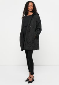 Abrigo largo negro acolchado con capucha, cierre de cremallera y bolsillos laterales, que se lleva sobre un top negro y pantalones ajustados negros. Zapatos de tacón negros.