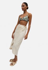 Top de bikini verde y blanco con estampado, falda envolvente beige con detalle de nudo y sandalias deslizables color tostado con diseño de nudo.