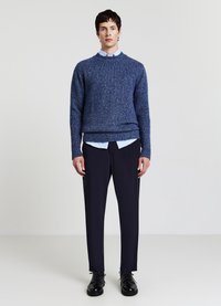 Pull texturé bleu porté sur une chemise à col bleu clair, assorti à un pantalon foncé et des chaussures noires à lacets, debout sur un fond clair.