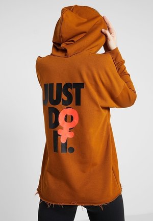 Person trägt einen burnt orange Hoodie mit dem Text "JUST DO IT" und einem pinkfarbenen weiblichen Geschlechtssymbol, das über dem "O" auf der Rückseite gesprüht ist.