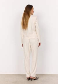 Completo sartorial color crema, caratterizzato da un blazer aderente con schiena strutturata e pantaloni a gamba ampia. Sono visibili un paio di décolleté a righe.