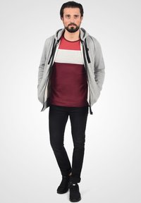 Grå zip-up hettegenser over en rød og grå stripete t-skjorte, kombinert med svarte jeans og svarte sneakers. En uformell, moderne antrekk med et lagdelt utseende.