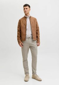 Mężczyzna w beżowej zamszowej kurtce, białej koszuli na guziki, jasnobeżowych spodniach i beżowych butach na sznurówki stoi na tle jednolitej białej ściany.