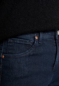 Mörkblå jeans i denim med en slät textur, med en framficka, kopparfärgade detaljer och subtila sömnadsdetaljer.