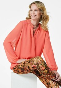 Korallefarbene Bluse mit Rüschenausschnitt und Schlüsselöffnungsdetail, kombiniert mit floral gemusterten, mehrfarbigen Hosen aus weichem, fließendem Stoff.