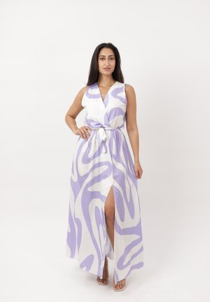 Mujer de pie, vestida con un vestido maxi blanco sin mangas con patrones abstractos en lavanda y una abertura frontal, posando con la mano en la cadera.