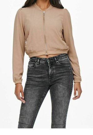 Vrouw draagt een beige cropped jas met rits en hoog getailleerde vervaagde zwarte skinny jeans tegen een witte achtergrond.