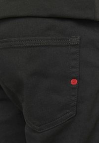 Los vaqueros de mezclilla negra tienen un bolsillo trasero con costuras visibles y un pequeño círculo rojo bordado como acento. La tela parece suave.
