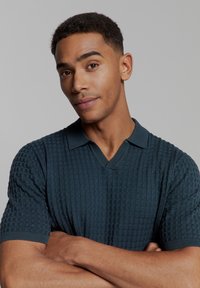 Gebreide polo shirt in donker teal met een gestructureerd patroon, korte mouwen en een V-halsontwerp. Zachte stof met een comfortabele pasvorm.