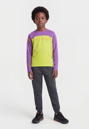 Junge mit kurzen lockigen Haaren, der ein lilafarbenes und gelbes Langarmshirt, dunkle Hosen und schwarze Turnschuhe mit orangenen Details trägt und steht.