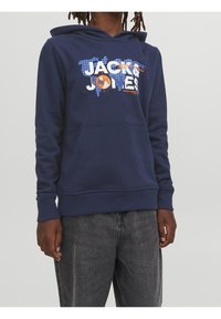 Námořnická modrá mikina s kapucí a přední kapsou, bílé a oranžové nápisy "Jack & Jones" nad modrou mapou světa, nošena s vybledlými černými džíny.
