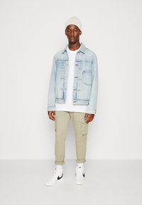 Chaqueta de mezclilla azul clara, camiseta blanca, pantalones cargo beige, zapatillas blancas con acentos negros y un gorro de punto beige claro.