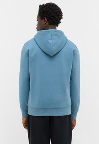 Mężczyzna z krótkimi kręconymi włosami, ubrany w niebieską bluzę i czarne spodnie, stojący tyłem na jednolitym jasnym tle.