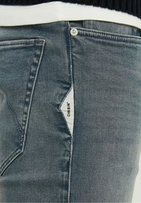 Jeans in denim blu scuro con una vestibilità slim, caratterizzati da una tasca angolata distintiva con un'etichetta tessuta e leggeri segni di usura sul tessuto.