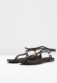 Black flat sandals with braided straps and gold accents, featuring ankle buckle closures, displayed on a white surface.