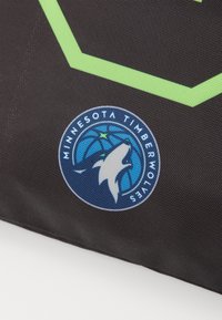 Sorte tekstil med et fremtrædende rundt logo, der viser en ulvehovede og en basketball, omgivet af blå og hvide accenter. Grøn geometrisk mønster inkluderet.