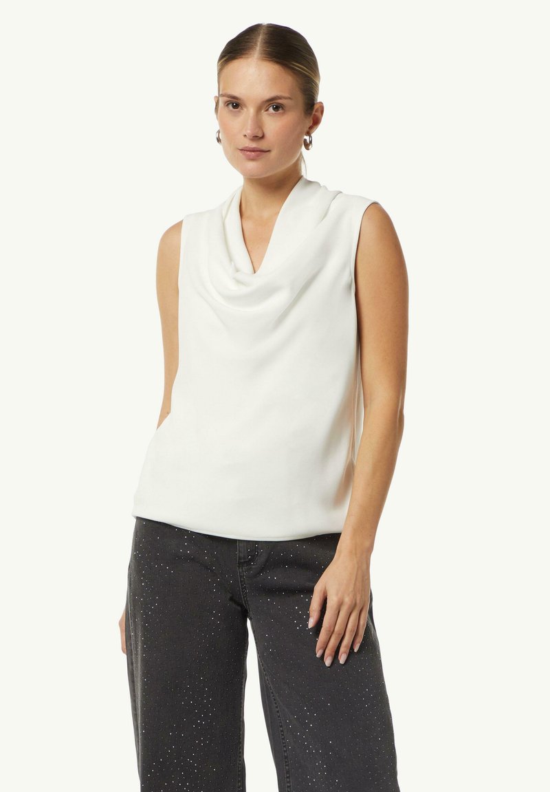 Biała bezrękawnik z drapowanym dekoltem, wykonana z gładkiego materiału; zestawiona z ciemnoszarymi dżinsami z srebrnymi ćwiekami.