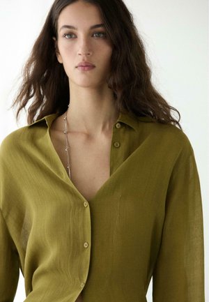 Donna con lunghi capelli castani, indossa una camicia verde oliva con bottoni e una delicata collana d'argento, in piedi davanti a uno sfondo bianco uniforme.