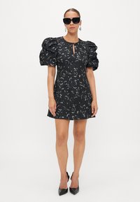 Čierne kvetinové šaty s nafúknutými rukávmi, detailom na krku vo forme kľúčovej dierky a textúrovanou tkaninou; doplnené čiernymi slnečnými okuliarmi a špicatými topánkami.