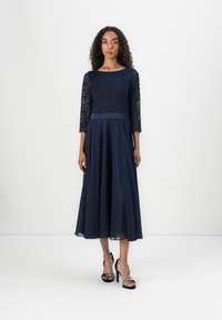 Robe marine avec un corsage en dentelle, des manches trois-quarts et une jupe en chiffon fluide, complétée par des talons hauts noirs à lanières.