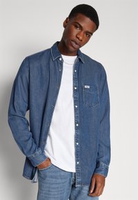 Chemise en denim bleu à manches longues, poche avant, fermeture à boutons, et un sous-vêtement bleu clair, assortie avec un jean bleu.