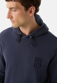 Muž hledící vlevo, oblečený v tmavě modré mikině s nastavitelnými šňůrkami a texturovaným logem "B" na hrudi.