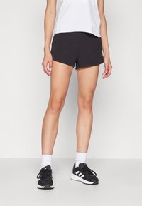 Shorts de sport noirs au design ajusté, en tissu léger, avec une petite fente sur le côté. Associés à un haut blanc et des chaussures de course noires.
