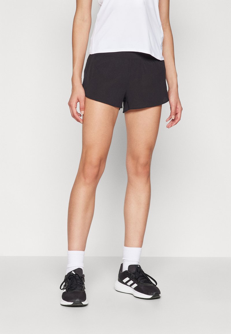 Shorts de sport noirs au design ajusté, en tissu léger, avec une petite fente sur le côté. Associés à un haut blanc et des chaussures de course noires.