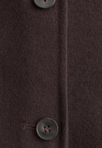 Tissu en laine marron foncé avec deux grands boutons noirs cousus, montrant des boutonnières et une couture verticale au milieu.