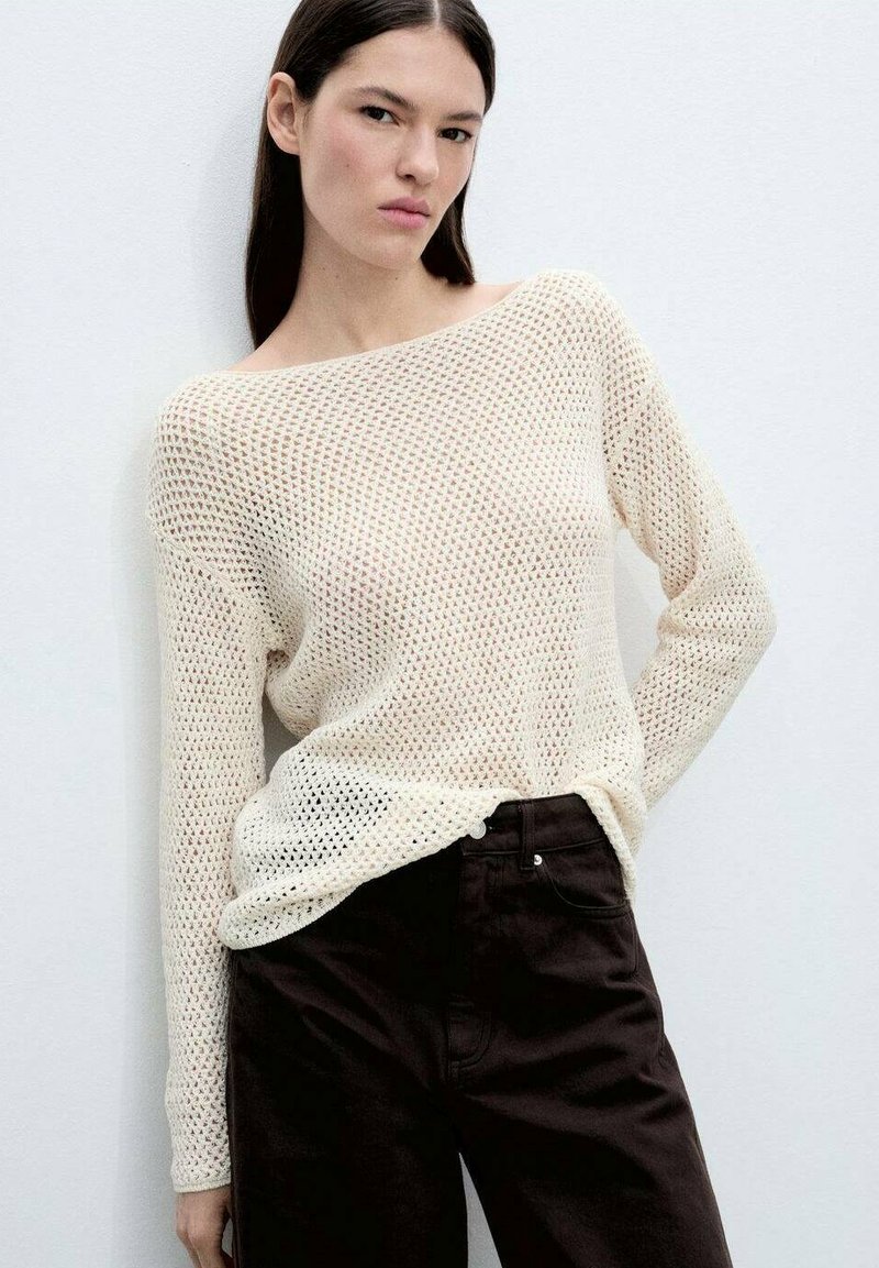 Helles Cremefarbenes Strickpullover mit offenem Gewebe, weitem, schulterfreien Ausschnitt und langen Ärmeln, kombiniert mit dunkelbrownem, figurbetontem Hosen.