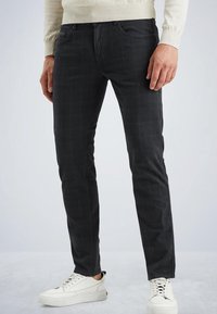 Man med slim-fit svarta rutiga byxor och vita sneakers, som står mot en ljusgrå bakgrund.