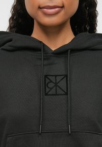 Μαύρη φούτερ με καπάκι που έχει μπροστινή τσέπη, κορδόνια και υφασμένο λογότυπο στο κέντρο, κατασκευασμένη από μαλακό ύφασμα με λεία υφή.