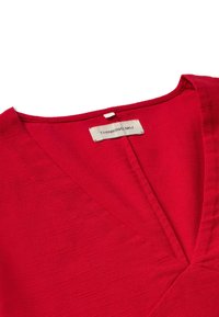 Top rojo con escote en V profundo, hecho de un tejido texturizado. La etiqueta visible dice "THINKING MU". Presenta líneas limpias y un acabado suave.