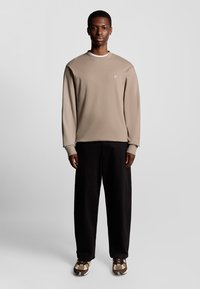 Beige sweatshirt med rund hals og subtilt logo, parret med løse sorte bukser og mønstrede brune sneakers.