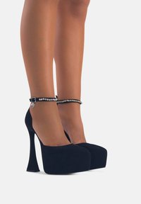 Tacco piatto nero realizzato in tessuto morbido, con tacco a spillo e un cinturino alla caviglia decorato con gioielli e un accento argentato.