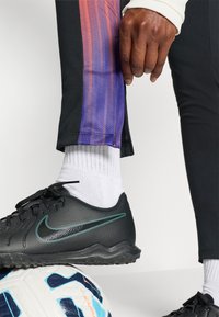 Pantofii sport negri cu accente verzi se află lângă o minge de fotbal. Pantalonii negri au un model gradient în nuanțe de violet. Sosetele albe sunt vizibile deasupra pantofului.