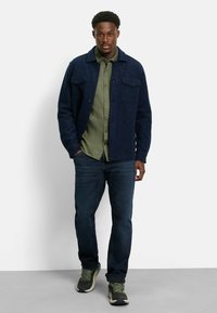 Mann in einer dunkelblauen Knopfjacke über einem olivgrünen Hemd, dunklen Jeans und grünen Turnschuhen, stehend vor einem schlichten Hintergrund.