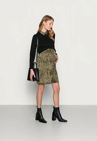 Mulher grávida vestindo uma blusa preta de mangas longas, saia de estampa de leopardo, botas pretas de tornozelo e segurando uma bolsa preta, de pé contra um fundo liso.