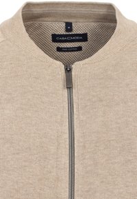 Beige zip-up cardigan lavet af Pima-bomuld. Har en struktureret indvendig side, rund krave og en sølvfarvet lynlås. Størrelse M mærkat synligt.