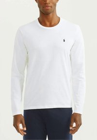 Άντρας που φοράει ένα λευκό μακρυμάνικο μπλουζάκι τύπου crewneck με ένα μικρό μαύρο λογότυπο παίκτη polo στο αριστερό στήθος και σκούρα παντελόνια.